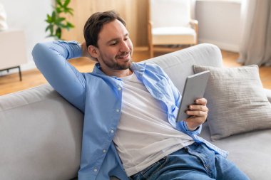 Bir adam tesadüfen bir kanepede oturuyor, tablet bir cihaz kullanıyor. Ekranla etkileşime girerken odaklanmış görünüyor. Günlük hayatta modern teknolojiyi sergiliyor..