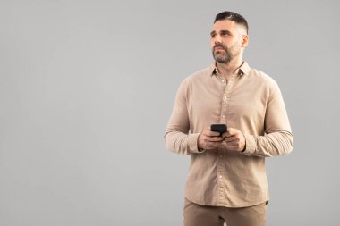 Bir adam elinde cep telefonu tutarken ayakta duruyor. Cihaza odaklanmış görünüyor, muhtemelen mesaj atıyor, arıyor ya da tarıyor. Arkaplan tanımsız, boşluğu kopyala