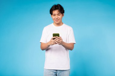 Beyaz tişörtlü Asyalı adam cep telefonuna dalmış, ekrana odaklanıyor. Cihazın üzerinde ciddi bir ifadeyle bir şey okuyor ya da kaydırıyor gibi görünüyor.