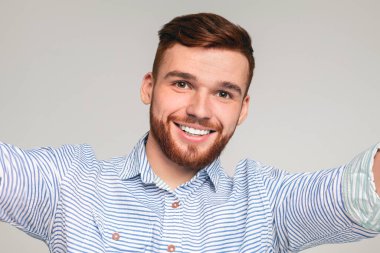 Yakışıklı milenyum sakallı adam özçekim yapıyor ve gri stüdyo arka planında gülümsüyor, kameraya gülümsüyor.