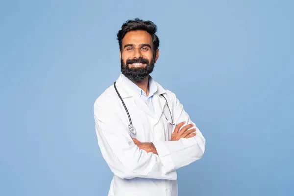 stock image A confident Indian doctor is smiling warmly, dressed in a white lab coat with a stethoscope draped around his neck, standing against a plain blue background.