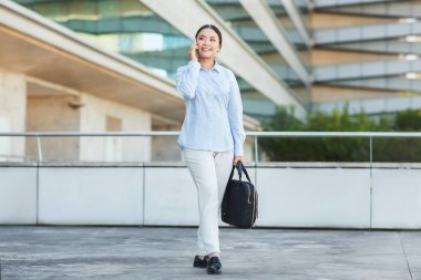 Asyalı bir iş kadını profesyonel bir ortamda telefonda konuşurken görülüyor. Çok yönlü yeteneklerini sergileyerek, konuya odaklanmış ve meşgul görünüyor..