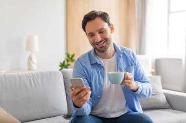 Bir adam rahat bir şekilde koltukta oturuyor, bir elinde bir fincan kahve, diğerinde de akıllı telefon kullanıyor. Adam rahat ve telefonuna odaklanmış görünüyor..
