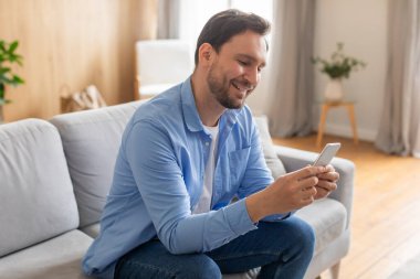 Kanepede oturan bir adam, telefon ekranına sabitlenmiş. Cihazında gösterilen dijital içeriğe dalmış gibi görünüyor..