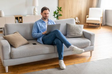 Bir adam kanepeye oturmuş, dizüstü bilgisayarına odaklanmış. Ciddi bir ifadeyle çevrimiçi olarak işine ya da aktivitelerine dalmış görünüyor. Oda basit, başka görünür kimse yok.
