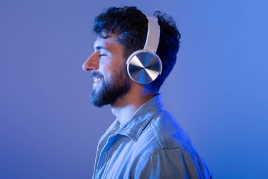 Sakallı bir adam kulaklık takıyor, müzik dinlemeye dalmış. Yüzünde ciddi bir ifadeyle sese odaklanmış görünüyor..