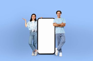 Bir adam ve bir kadın boş bir ekranın yanında durup resim için poz veriyorlar. Kadın beyaz bluz ve siyah pantolon giyerken adam siyah takım elbise giyiyor..