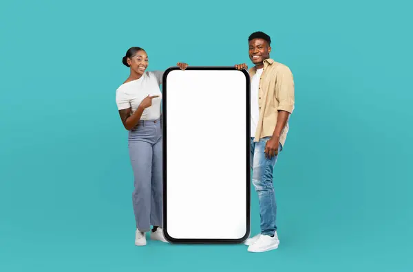 stock image Black man and woman are standing against a blue background, holding up a blank screen. They appear to be showcasing something on the screen, potentially for a presentation or advertisement.