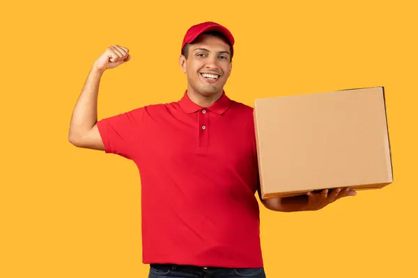 stock image A cheerful delivery worker man in a red cap and polo shirt holds a cardboard box while flexing his arm, showing enthusiasm and strength.