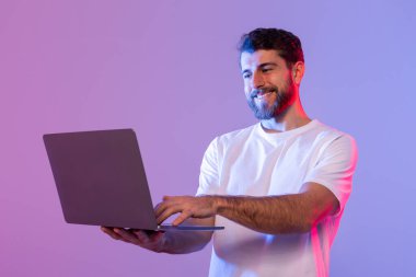 Beyaz gömlekli bir adam ayakta duruyor ve elinde bir dizüstü bilgisayar tutuyor. Ekrana odaklanmış görünüyor, muhtemelen çalışıyor ya da çalışıyor. Arka plan basit ve dikkat dağıtıcı unsurlardan yoksun..