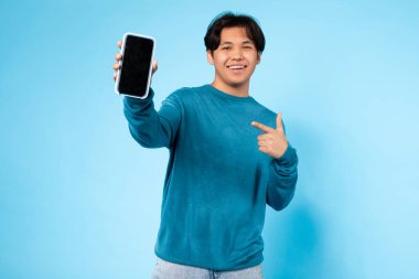 Mavi kazak giyen Asyalı genç adam diğer eliyle akıllı telefonu göstermekten mutluluk duyar. Katı mavi bir arkaplanda duruyor.