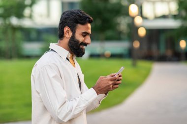 Sakallı bir Hintli cep telefonu ekranına odaklanmış, muhtemelen okuyor ya da mesaj yazıyor. Hareketsiz dururken dikkati cihaz tarafından tamamen çekiliyor..