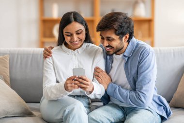 Hintli bir erkek ve kadın koltukta oturuyor, ikisi de dikkatle cep telefonu ekranına bakıyor. Yüzlerinde odaklanmış ifadelerle cihazda gösterilen şeylerle meşgul görünüyorlar..