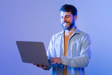 Mavi gömlekli bir adam oturuyor ve aktif olarak dizüstü bilgisayar kullanıyor. Elleri klavyenin üzerinde ve gözleri çalışırken ekrana odaklanmış durumda..