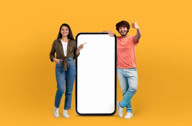 Hintli bir adam ve kadın birlikte duruyorlar, boş bir telefon ekranını işaret ediyorlar. Odaklanmış ve meşgul görünüyorlar, muhtemelen içeriği tartışıyorlar veya cihaz üzerinde bir özellik gösteriyorlar.