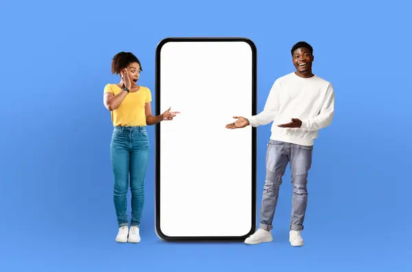 stock image A man and a woman are standing side by side, looking at a blank screen on a phone. The man is holding the phone, while the woman is gesturing towards it with a curious expression.