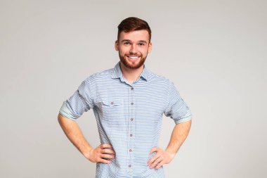 Gri stüdyo arka planında elleri kalçalarında, günlük tişört giyen gülümseyen adamın portresi.