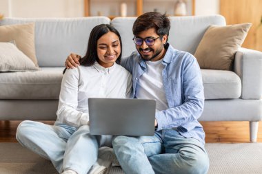 Hintli erkek ve kadın yerde oturuyorlar, her ikisi de önlerindeki dizüstü bilgisayara odaklanmış. Bilgisayarda gösterilen şeylerle meşgul görünüyorlar, birbirlerine doğru eğiliyorlar..