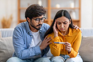 Genç bir Hintli çift gün boyunca oturma odasındaki kanepede birlikte oturur. Kadın hamilelik testi yaparken endişeli görünüyor, ve ortağı da rahatlatıcı destek öneriyor.