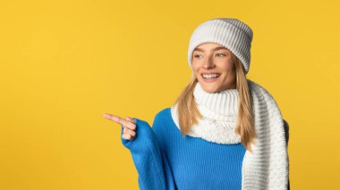 Mavi kazak, beyaz eşarp ve uyumlu örgü şapka giymiş neşeli genç bir kadın canlı sarı bir arkaplanda fotokopi uzayını göstererek kenarı işaret ediyor.