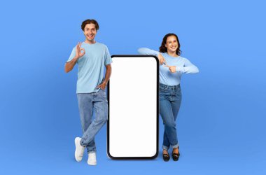 Gülümseyen adam ve kadın yan yana duruyorlar. Kadının elinde tuttuğu telefonun boş ekranına bakıyorlar. Adam ekranı işaret ediyor ya da işaret ediyor..