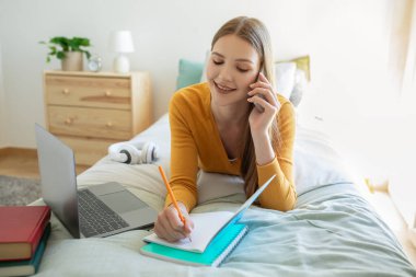 Kız sabahları yatağında uzanıyor, telefonla konuşuyor, defterine yazıyor, evde çalışıyor, dizüstü bilgisayarı ve kitapları yanında, verimli ve düzenli bir ortam olduğunu gösteriyor..