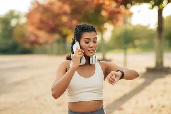 Stock image A young woman dressed in athletic clothing is in a park during the autumn season, talking on smartphone, with wireless headphones around her neck, while simultaneously glancing at her smartwatch.