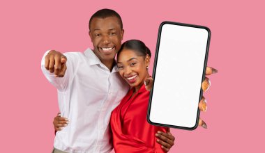 Mutlu bir siyah çift pembe bir arka planın önünde duruyor. Adam sol eliyle kamerayı işaret ediyor. Kadın elinde boş ekranlı bir akıllı telefon tutuyor.