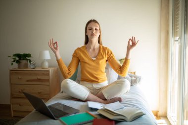 Genç bir kız yatağında oturmuş, gözleri kapalı ve sakin bir ifadeyle meditasyon yapıyor. Etrafı açık kitaplar, dizüstü bilgisayarlar ve defterlerle çevrili. Bu da çalışmaya ara verdiğini gösteriyor.
