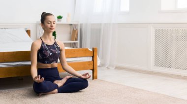 Genç bir kadın huzurlu bir yatak odası ortamında meditasyon yapıyor, bağdaş kurarak bir yatağın önünde oturuyor..