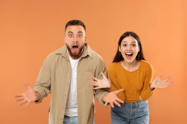 Genç bir çift birlikte duruyor, her ikisi de şaşkınlık ve heyecan ifadeleri sergiliyor, geniş gözler ve açık ağızlarla. Gelişigüzel giyinmişler ve arka planda katı bir portakal var.