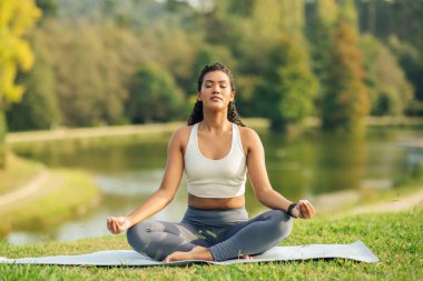 Yoga minderinde bağdaş kurmuş genç bir kadın yemyeşil bir parktaki dingin bir gölün kenarında meditasyon yapıyor. Sakin bir sabah. Rahatlamış ve odaklanmış görünüyor. Doğanın huzurunu kucaklıyor..