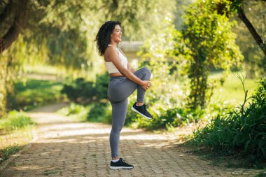 Genç bir kadın sabah yemyeşil bir parkta bacak esneme hareketleri yapıyor. Odaklanmış ve enerjik görünüyor, temiz havanın ve huzurlu çevrenin tadını çıkarıyor..