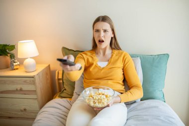 Bir kız elinde patlamış mısır kasesi ve uzaktan kumandayla yatakta oturuyor. Rahat bir ortamda televizyon izlemeye hazırlanırken rahatlamış görünüyor..