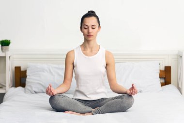Genç bir kadın bir yatakta meditasyon pozisyonunda oturuyor, gözleri kapalı, aydınlık, aydınlık bir odada..
