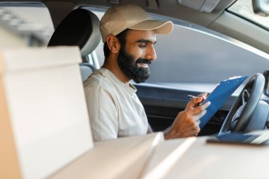 Hintli kurye aracının sürücü koltuğunda otururken teslimat listesini kontrol ediyor. Aracın içinde birkaç paket görünüyor. Bu da yoğun bir teslimat programı olduğunu gösteriyor..