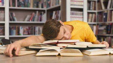 Kütüphanede uyuyor. Yorgun beyaz adam kitap yığınında şekerleme yapıyor, sınavlara hazırlanırken yoruluyor