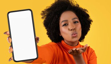 Genç siyahi bir kadın önünde akıllı telefon tutuyor ve öpücük yolluyor. Sağ eli telefonu tutarken, sol eli bir öpücük şeklinde ve izleyiciyi işaret ediyor.