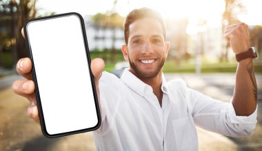 Beyaz gömlek giyen genç bir adam gün ışığında dışarıda duruyor. Elinde boş bir ekranla akıllı bir telefon tutuyor, gülümsüyor ve doğrudan kameraya bakıyor..