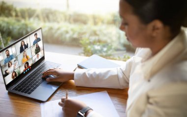 Genç bir kadın, gün içinde bilgisayarında ofis masasında oturmuş, kalemle notlar alırken ve çalışma alanının etrafına dizilmiş not defterleri varken sanal bir takım toplantısına katılıyor.