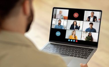 Bir grup insan ekranda görünürken bir dizüstü bilgisayar kullanan bir adam. Kişi, laptop ekranında gösterilen kişilerle sanal iletişim ya da işbirliği içindedir..