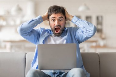 Kanepede oturan bir adam, dizüstü bilgisayarına şok içinde bakıyor. Elleri başının üstünde ve ağzı şaşkınlıkla açık. Ekranda gördüğü bir şeye tepki veriyor gibi görünüyor.