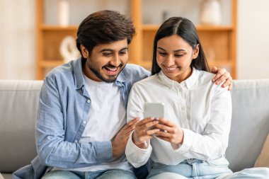 Kanepede oturan Hintli çift, ikisi de cep telefonu ekranına bakmaya odaklanmış. Konsantrasyon ve ilgi ifadeleri ile cihazda gösterilen her şeyle ilgili görünürler..