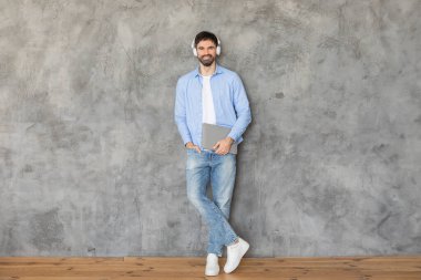 Bir adam beton bir duvarın önünde duruyor, bir eli cebinde duvara yaslanıyor. Kameraya gülümsüyor ve kulaklıklarının üzerine kulaklık takıyor. Diğer elinde bir dizüstü bilgisayar tutuyor.