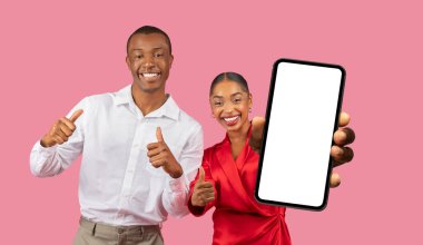 Bu görüntüde mutlu bir Afro-Amerikalı çift pembe bir arka planın önünde duruyor. Kadın, önünde boş bir ekranla akıllı bir telefon tutarken, erkek baş parmağını kaldırıyor.