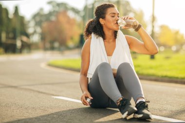Bir kadın yol kenarına oturmuş, ağzına bir şişe dayamış ve ondan içiyor. Rahatlamış ve içkisine odaklanmış görünüyor..