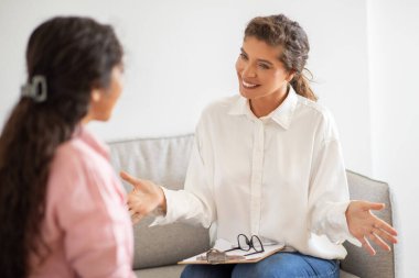 Beyaz gömlekli bir kadın kanepede oturuyor ve bir müşteriyle konuşuyor. O, elleriyle hareket ediyor ve gülümsüyor. Müşteri dikkatle dinliyor. Bu resim tipik bir terapi seansını betimler.