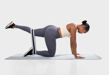 Beyaz spor sutyen ve gri taytlı Afro-Amerikalı kadın mavi yoga minderi üzerinde glute briç egzersizi yapıyor. Kalçasının etrafında siyah-beyaz direniş bandı kullanıyor.