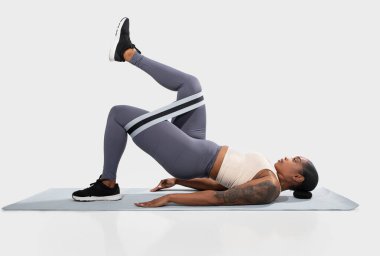 Atletik kıyafetli Afrikalı Amerikalı bir kadın yoga minderinde bir direniş bandıyla köprü egzersizi yapıyor. Dizleri bükülmüş ve ayakları paspasın üzerinde sırt üstü yatıyor..