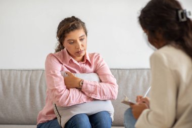 Pembe giyinmiş genç bir kadın, ofis ortamında bir koltukta oturuyor, göğsüne yastık dayamış, aşağıya bakıyor. Zor durumda gibi görünüyor..